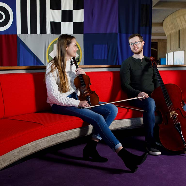 A student with a violin and another with a cello sit on a couch and talk.